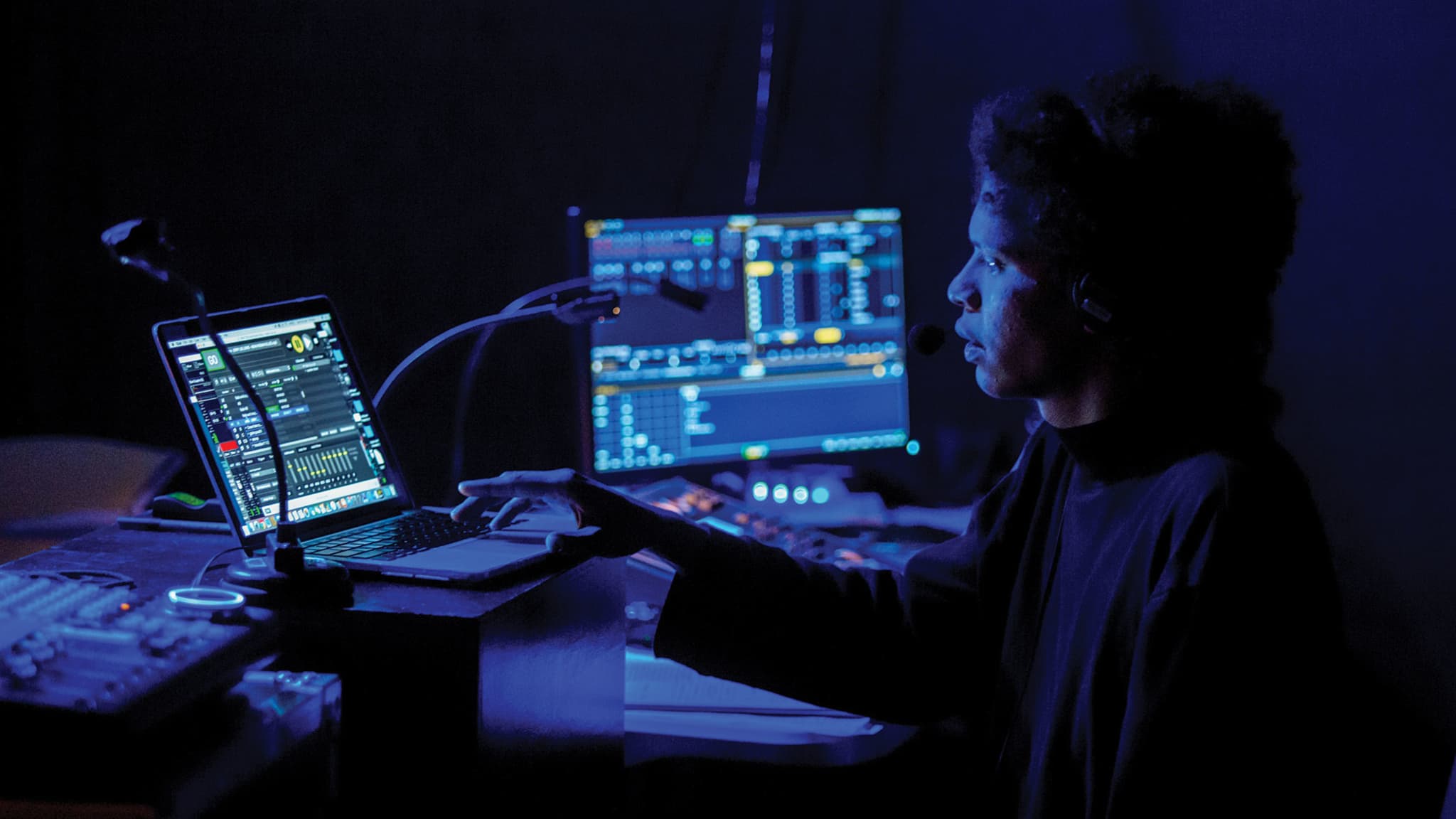 A student sits in the dark behind two computers, a mic, and sound and lighting boards. They are illuminated by the blue light of their screens.