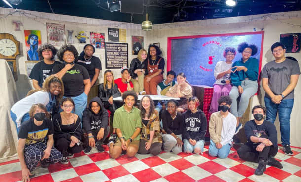 A group of students gather together on a small stage with white and checkered flooring. Some sit on the ground, some stand, and some sit in a corner booth at the center of the stage.