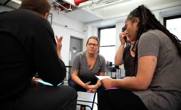 A small group of adults in black folding chairs sit in a circle in a rehearsal studio and have a discussion.