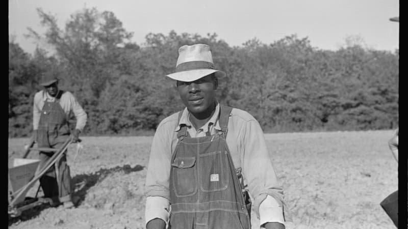 A dark-skinned man in overalls, a collared shirt, and low-brimmed hat in a farm field. In the background is a similarly dressed dark-skinned man pushing a farm implement.