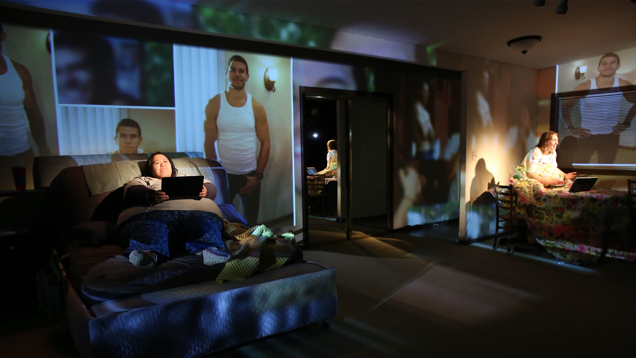 Production photo from KINGDOM COME. A plus-sized person sits on a couch with a laptop, and its light illuminates her face. Images are projected behind her.