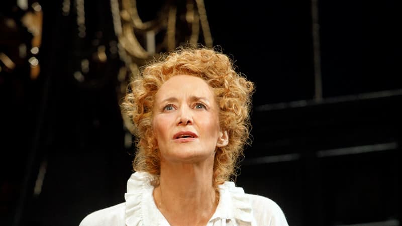 A woman with very short curly blonde hair wears a white Shakespearean blouse with a frilly collar and black trousers.