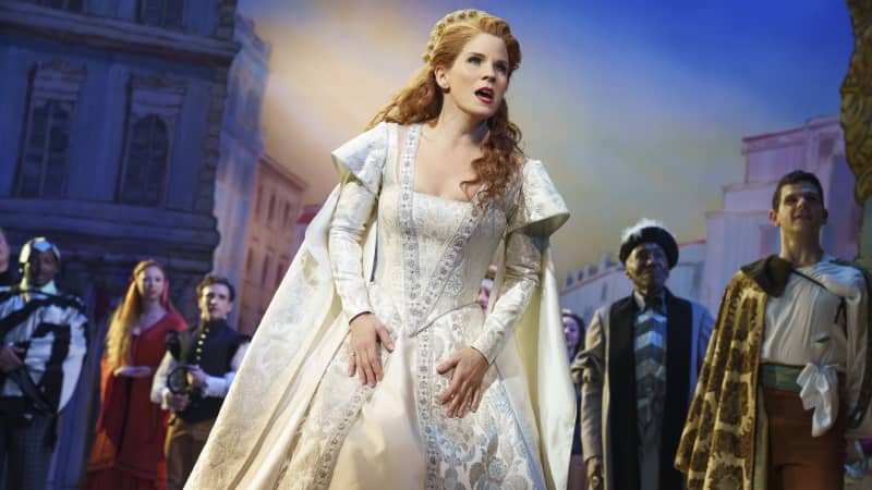 A woman with red curls wears a white, royal Shakespearean dress on stage with a Shakespearean ensemble behind her.