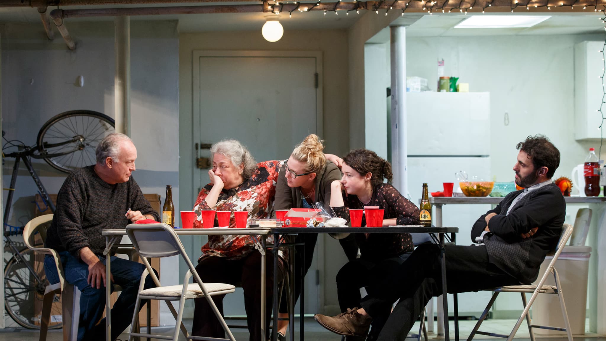 Five actors on stage sit around two card tables pushed together and littered with red solo cups and beers. From THE HUMANS.