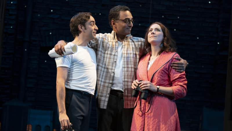 Two men and a woman stand together with arms around each other. Two of them hold binoculars, and the woman is in a night robe and looks at a starry night city sky.