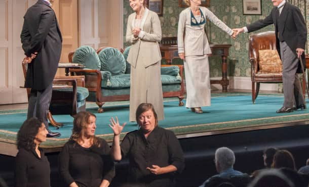 Three people dressed in black stand in front of a stage using sign language while a play performs behind them.