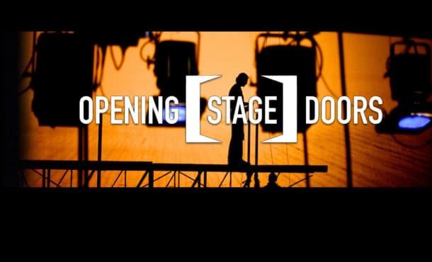 Opening Stage Doors. A silhouette of a person standing on a platform surrounded by theatre lights. The background silhouetting them is orange.