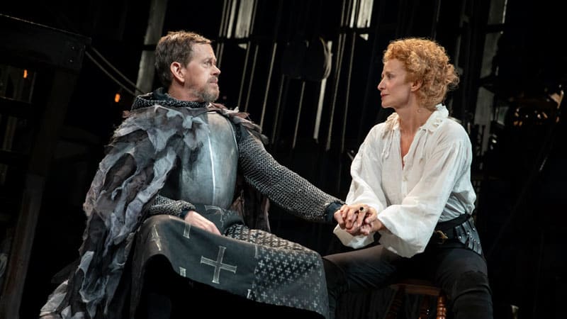 A woman with very short curly blonde hair in a white Shakespearean blouse and black trousers sits and compassionately clasps the hand of a man dressed as a Shakespearean knight.