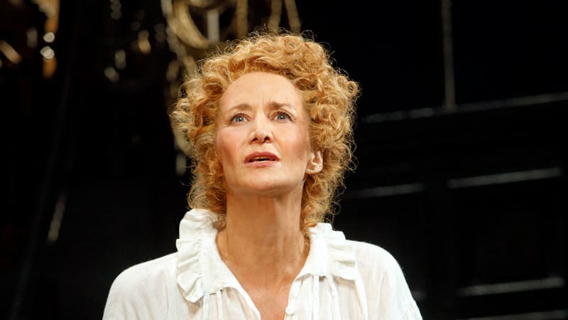 A woman with very short curly blonde hair wears a white Shakespearean blouse with a frilly collar and black trousers.