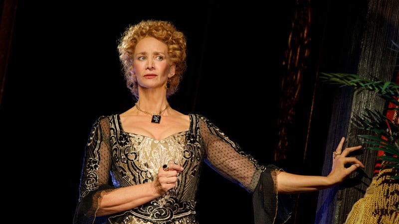 A woman with very short curly blonde hair wears a fancy gold and black Shakespearean dress and a square, black pendant necklace.