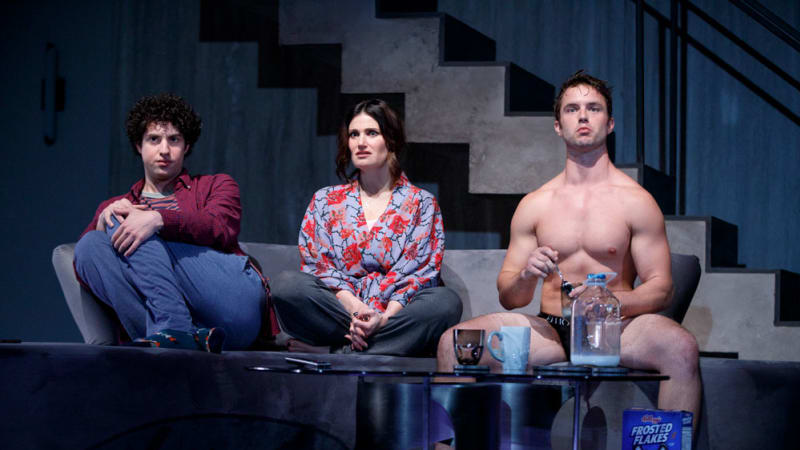 A young man and a woman sit with their legs tucked on a sofa next to a shirtless, muscular man in his underwear as they all watch TV.