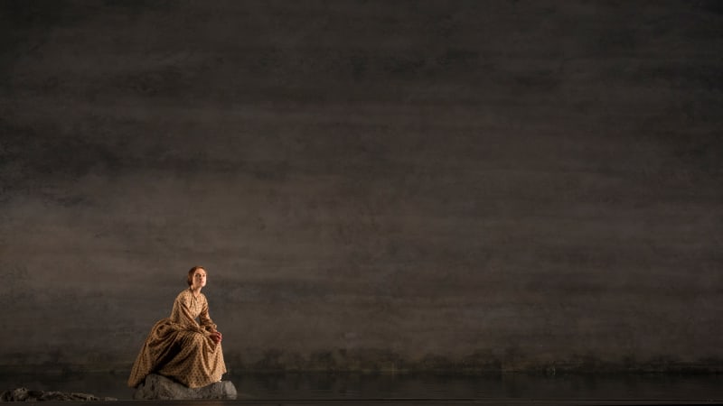 On a large, vast gray stage, a woman in a low bun and an 1860s style brown dress crouches on a small boulder. She looks out into the distance.