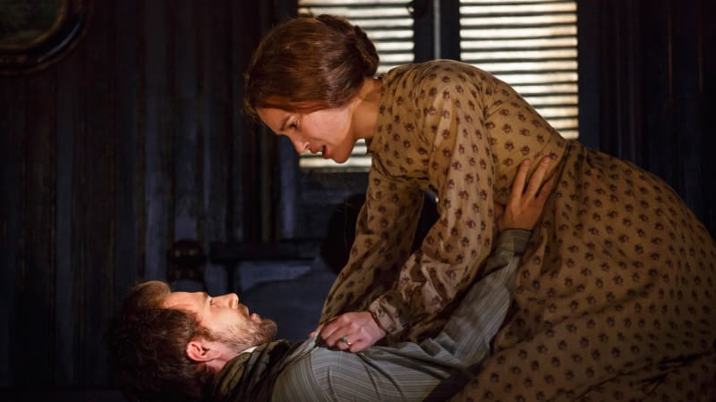 A woman kneels over a man who has fallen back towards the ground and grips his shirt with worry. She wears an 1860s brown dress and low bun. He wears a best over a striped shirt.