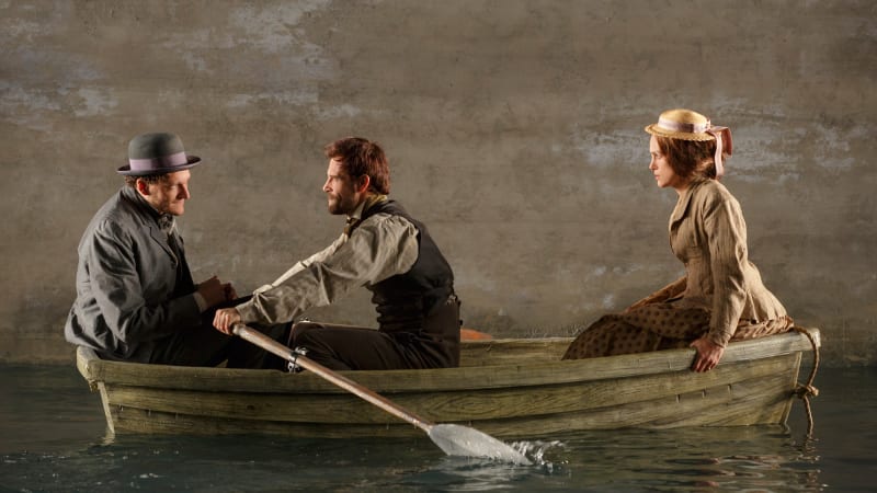 A man and woman in 1860s clothing sit on either ends of a wooden boat, while a man in the middle paddles them over a body of water.