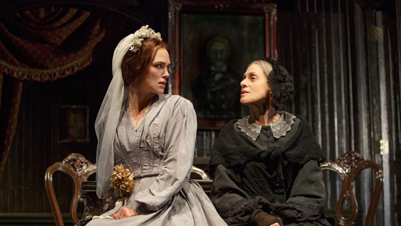 A woman in a gray 1860s dress and wedding veil sits next to a woman in a black 1860s gown and headpiece. The conversation seems tense.