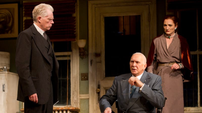 One man in a suit sitting down while another man is standing over him and woman is behind them both. 