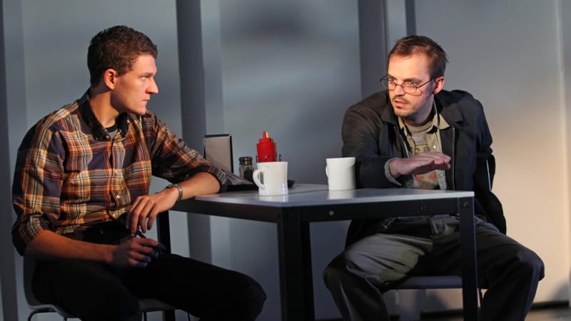 Two men sitting at a diner table both looking fairly concerned. 