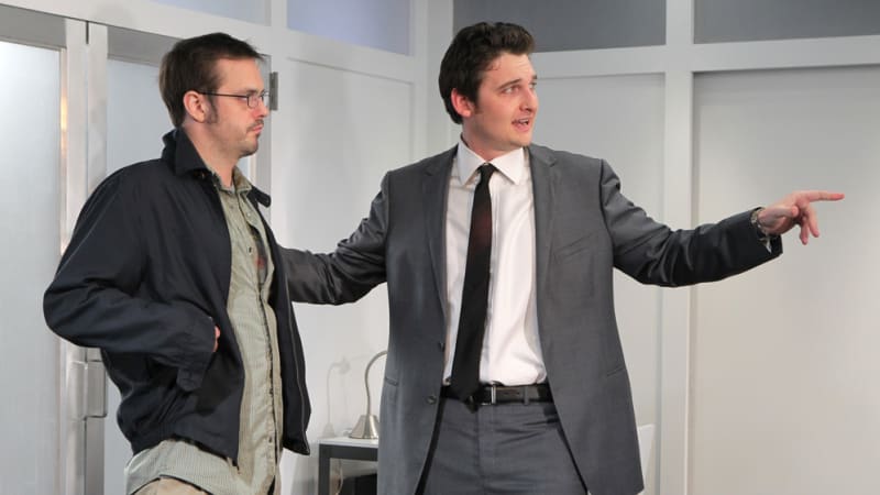 Two men in a room. One man wearing a gray suit has his hand on the others shoulder and is pointing something out. 