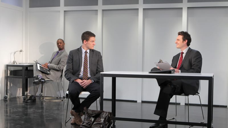 3 men in a room all wearing suits. It looks like an interview with one man smiliong and holding a clipboard. 