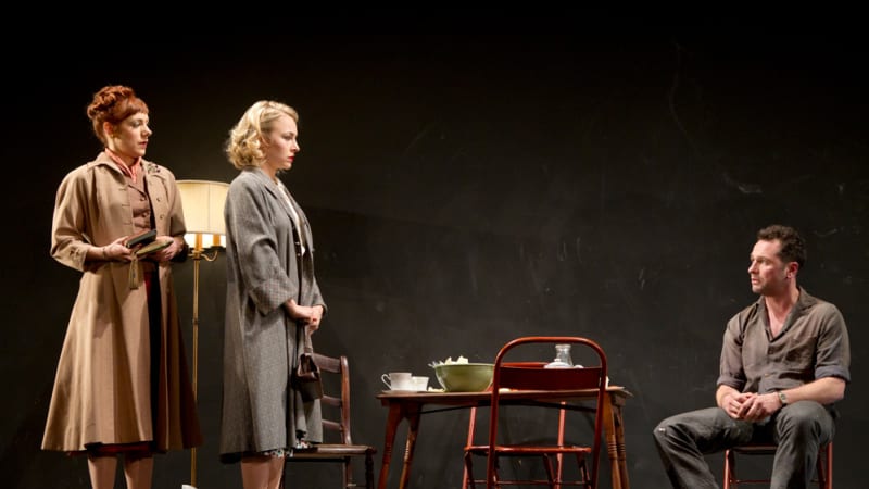 Two women in coast stand together in a dirty room over a man who seems sad sits in a chair and looks at them. 