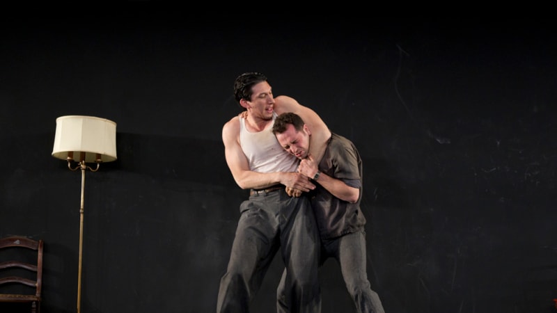 Two men fighting. A man in a white tank top has the other in a chokehold. The room is destroyed.