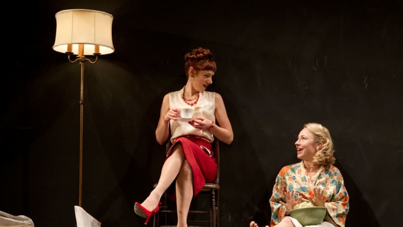 two women sitting and chatting in a messy room. One woman in a red skirt is sitting in a chair drinking a cup of coffee while the other lays on the floor. 