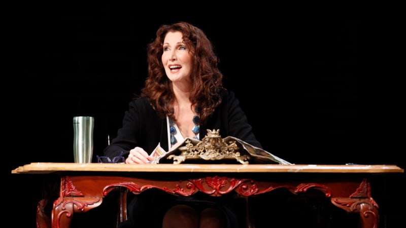 A woman smiles while sitting at an ornate desk reading a magazine. 