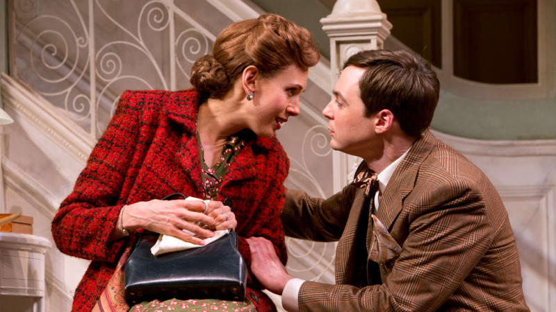 A woman in a red coat is sitting holding her purse and is leaning into kiss a man in a brown suit kneeling next to her. 