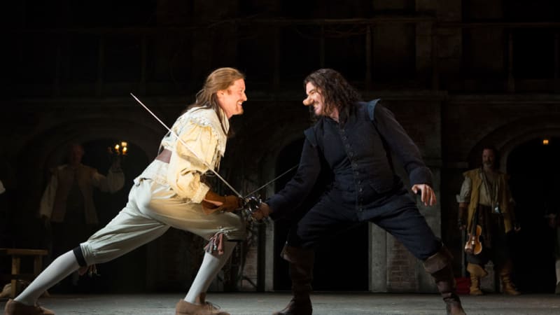 A man in dark 17th century clothes and a large nose has a sword fight with a blonde man in white.