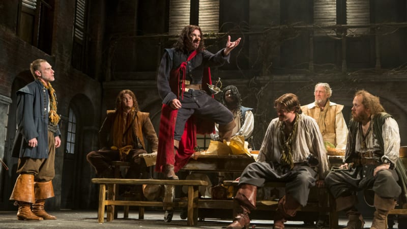 A man with a large nose and a red cape stands on a wooden lunch table and makes a proclamation to a group of other men in 17th century clothes.