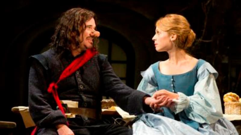A man with long brown hair and a large nose wears dark 17th century clothes with a red sash. He sits next to and holds the hands of a blonde woman in a blue dress and corset.