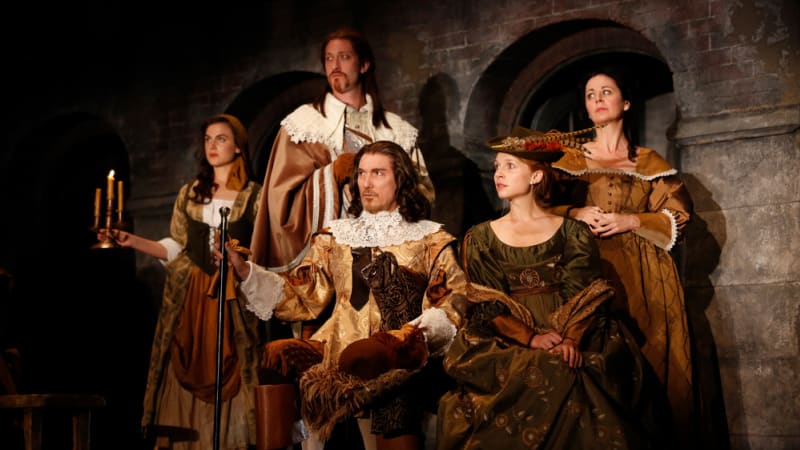 Four actors in 17th century clothing. One of them holds a small candelabra, two in slightly fancier clothes sit in front of the rest, one of them holding a staff.