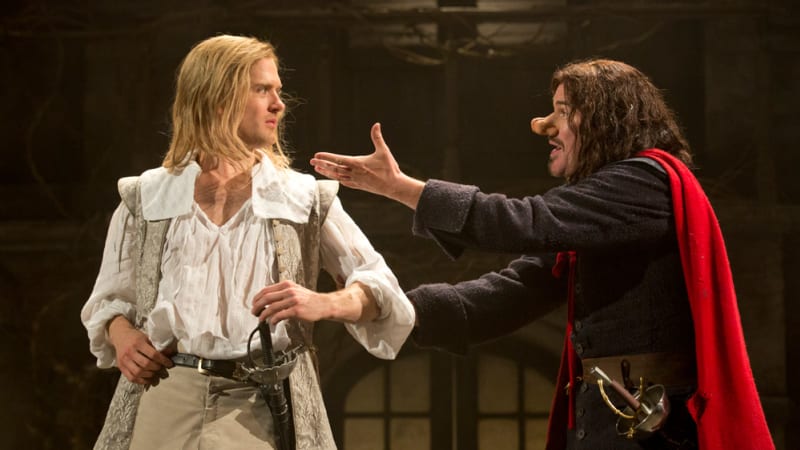 A man with long blonde hair and light-colored 17th century clothing rests a hand on his sheathed sword. A man with long hair, a large nose, and a red cape gestures towards him.