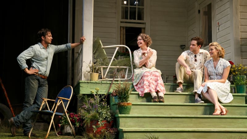Two women and one man happily sit on the green steps of a back porch, while a man in a tattered shirt and old jeans leans against the porch's column with a hand on his hips.