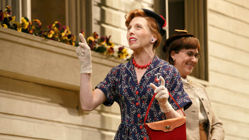 An older woman with red hair, a hat, and gloves holds a red purse and points up at a house. Behind her is another woman in similar clothing.