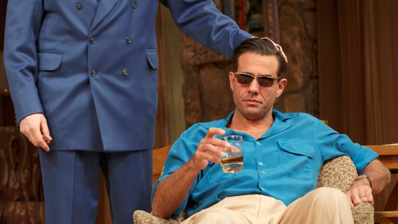 An older man in a blue double-breasted suit pats the head of a man in sunglasses holding a glass of liquor lounging in a fancy chair.