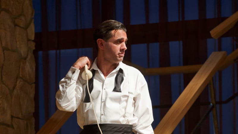 A man in a tuxedo without the blazer and an undone bowtie holds a rotary phone in one hand with the receiver up by his shoulder.