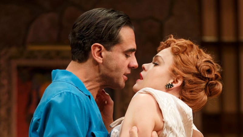 A man in a bright blue shirt and tan pants clutches the arms of a red-headed womanin white and pulls her close to his face.