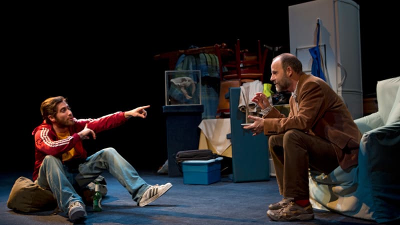 Two men are sitting in a room, one on the ground and one in a chair. The man on the ground is pointing at the man in the chair. 