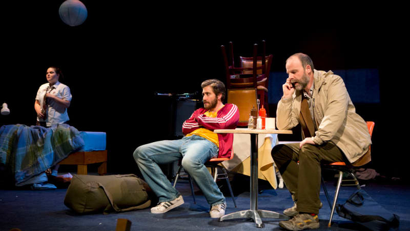 Two men are sitting at a table while one is on the phone. In the background there is a girl getting into a school uniform behind a bed. 