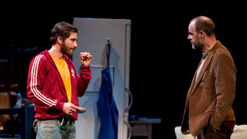 A man in a red addidas track jacket is smoking a cigarette while he looks at another man. 