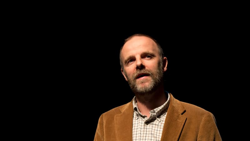 A man stands with his hand out in a spotlight wearing a corduroy tan jacket. 