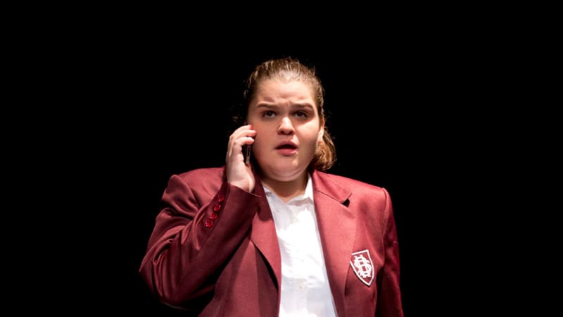 A girl in a red school uniform is leaning against a bath tub while holding a cellphone to her ear. 