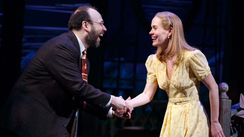 A man and a woman shake hands and laugh with each other. He has a short beard, glasses, and wears a suit. She has blonde hair and wears a yellow 1940s dress.