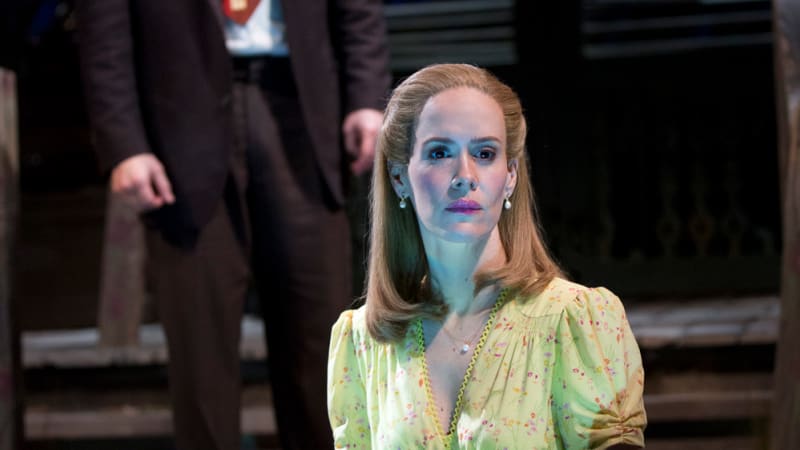 A blonde woman in a 1940s yellow dress sits at the end of the pier, blue water reflecting on her face as she stares out. A man with a beard wearing a suit stands behind her.