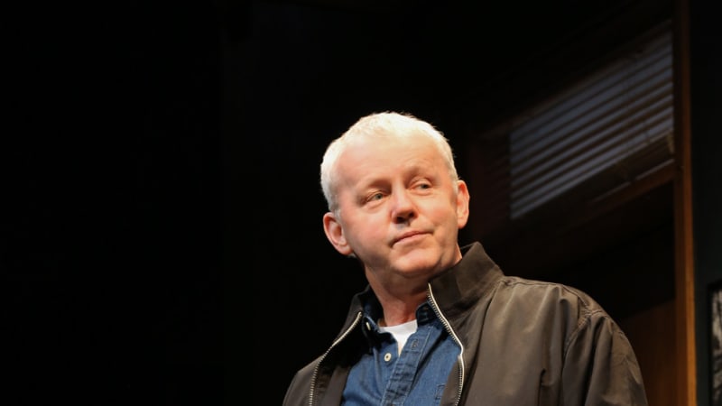 An older man wearing a black jacket over a denim shirt and tan pants. He puts one hand in his pocket, the other holding a black bag.