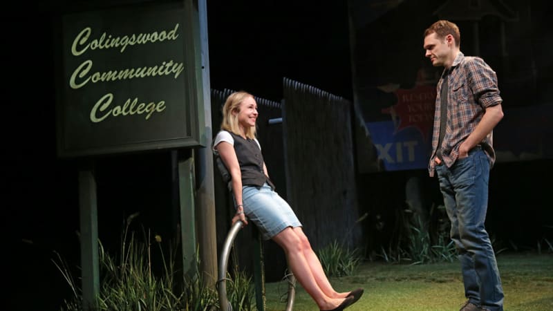 A young woman in a vest and skirt playfully leans against a bike rack by a sign that says Colingswood Community College. She talks with a young man in a flannel.
