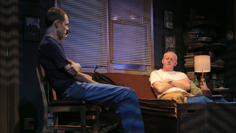 in a messy living room at night, with blinds casting shadows across the stage, an older man crosses his arms on the couch and speaks to a younger man who sits on a chair and holds his own arm.
