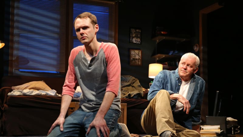 In a messy living room set, a young man kneels on the floor with a solemn expression, facing away from an older man who sits on the floor and looks at him.
