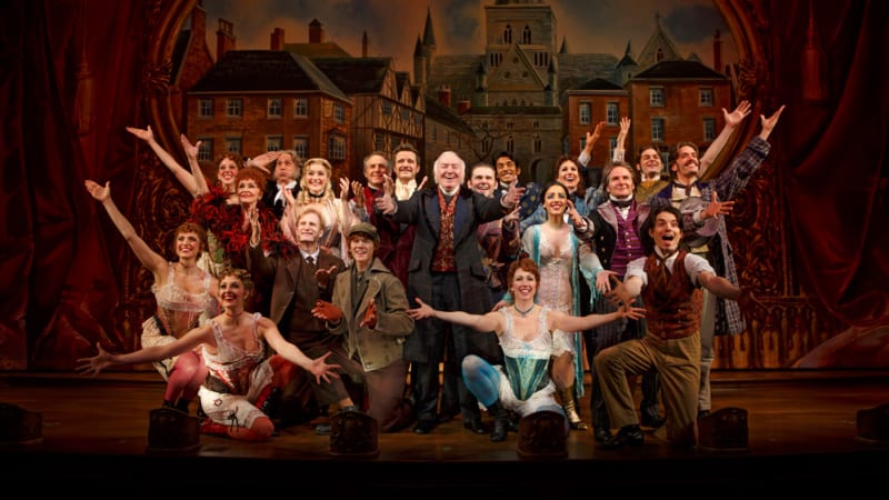 The entire cast, dressed in 1870s clothing, poses together in a clump with their arms outstretched. A painted background of an English town is behind them.
